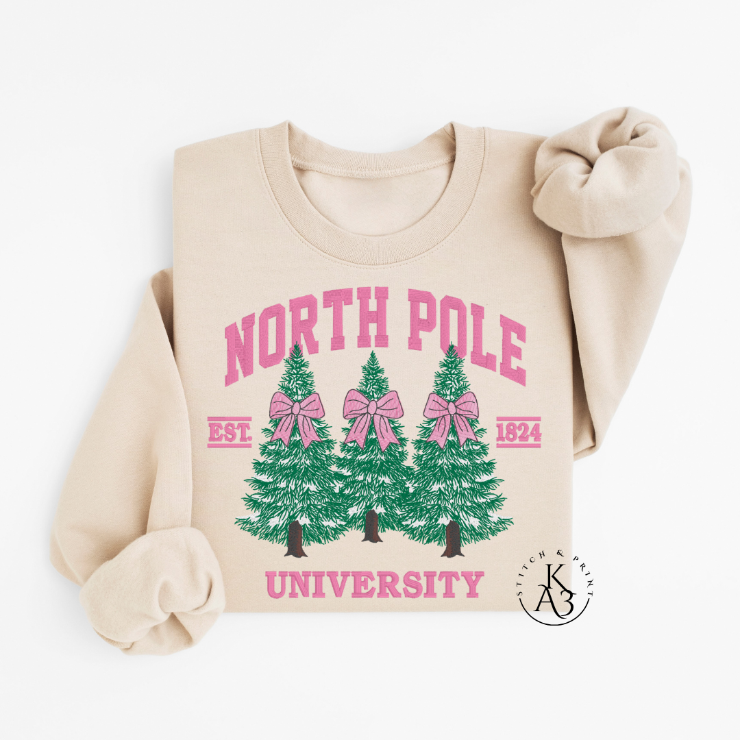 Beige sweatshirt with embroidered 'North Pole University' design featuring trees and bows on a white background.